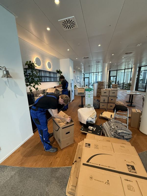 In einem Großraumbüro füllt ein Mitarbeiter Umzugskartons, auf dem Holzboden liegen zusammengelegte Kartonagen neben aufgestapelten Kisten