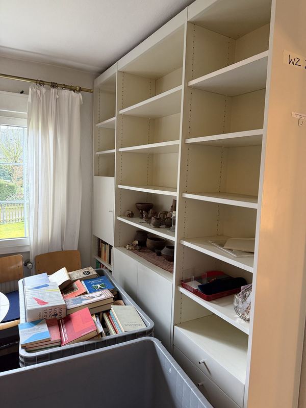 Das Bücherregal ist fast geleert Ein weißes Regal für Bücher, fast vollständig geleert, davor ein grauer Rollbehälter, in dem Bücher liegen