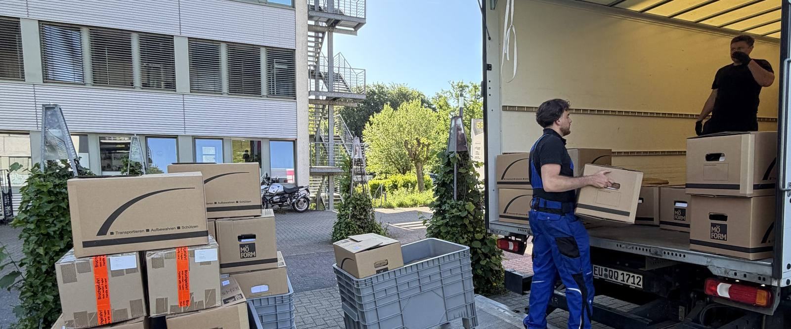 Einblicke in unseren Umzugsalltag in Bornheim: Vor einem Bürogebäude mit Außentreppe steht ein Umzugswagen mit offenem Laderaum, ein Mitarbeiter trägt einen Karton zur Ladebordwand, ein zweiter steht im Laderaum, im Vordergrund gestapelte Umzugskartons und graue Kunststoffboxen.