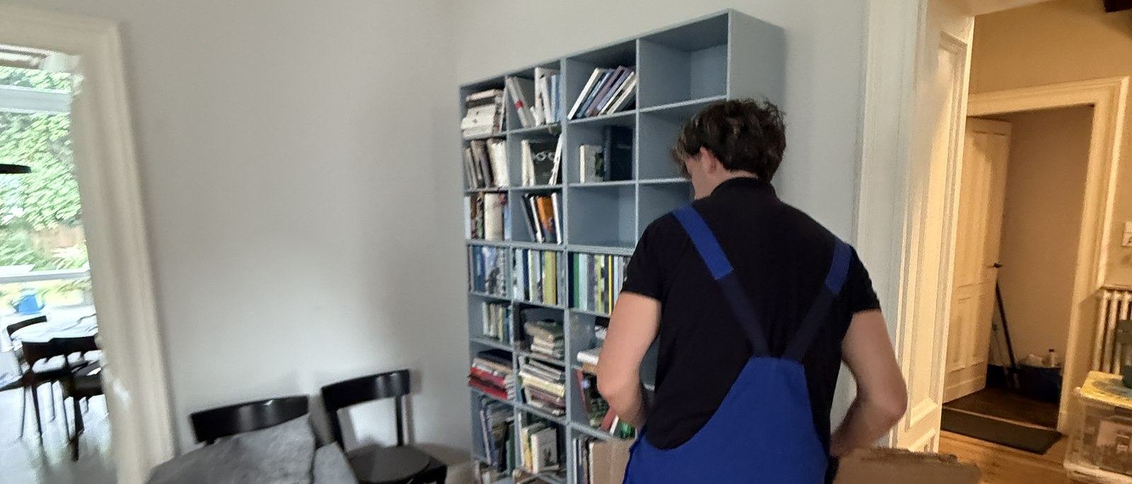 Fragen und Antworten zum Umzug in Bornheim: Ein Mitarbeiter in Latzhose steht vor einem hellgrauen Würfelregal mit Büchern und Ordnern und packt dessen Inhalt in einen Karton, im Vordergrund ein Esstisch mit Stühlen.