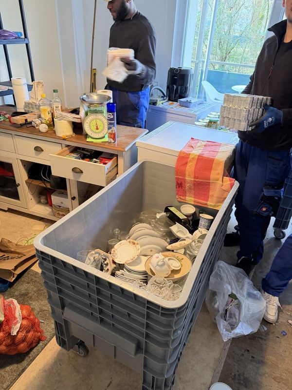 Geschirr und anderen Hausrat legen zwei Kollegen in einen großen grauen Rollcontainer