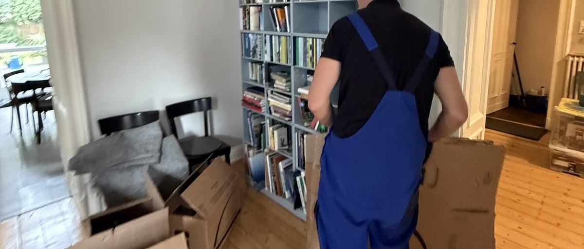 Haushaltsauflösung in Bornheim: Ein Umzugshelfer in Latzhose geht mit einem zusammengefalteten Karton an einem gefüllten Bücherregal entlang, im Vordergrund Stühle und noch mehr Kartons.