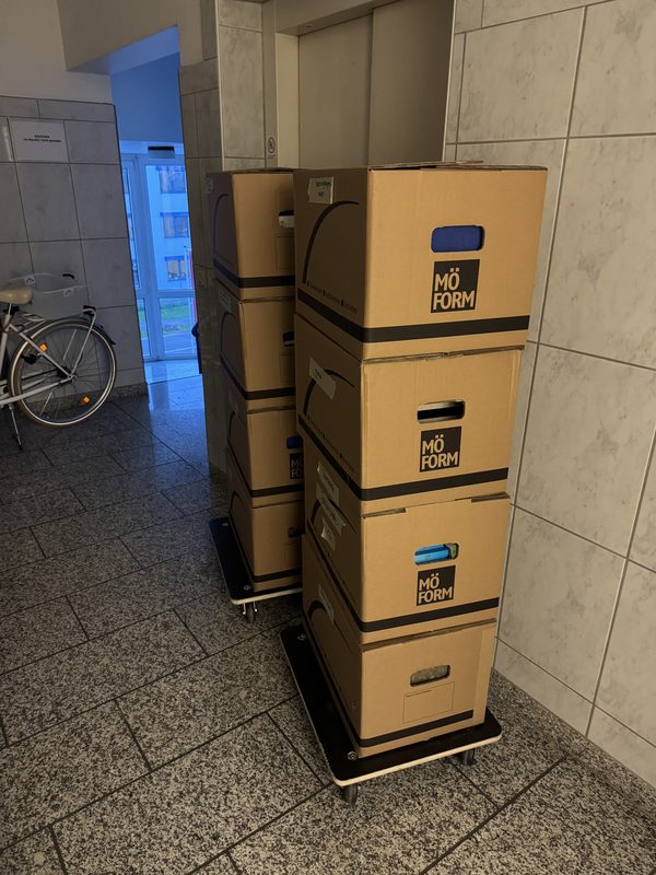 Auf einem Rollbrett stehen vier Kartons übereinander in einem gefliesten Flur, dahinter ein Fahrrad