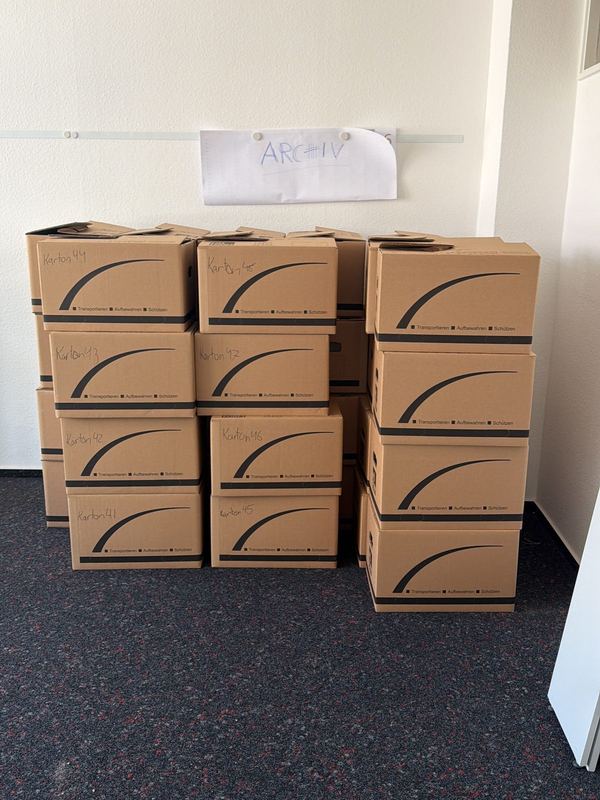 Kartons, nach Zielräumen sortiert Von Hand beschriftete Umzugskartons, gestapelt in einem leeren Zimmer mit dunklem Teppich, an der Wand klebt ein Zettel mit dem Wort Archiv