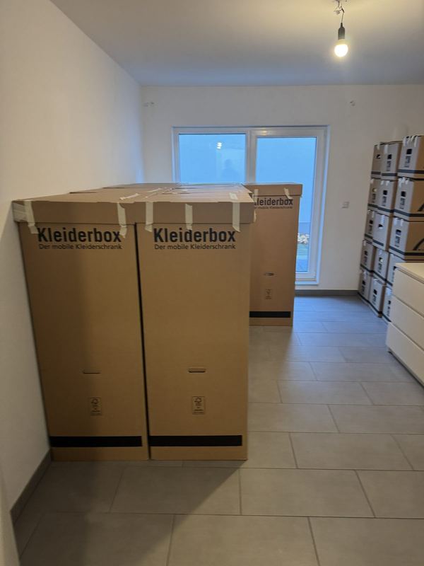 In einem leeren Raum stehen zwei Kleiderboxen neben gestapelten Kartons