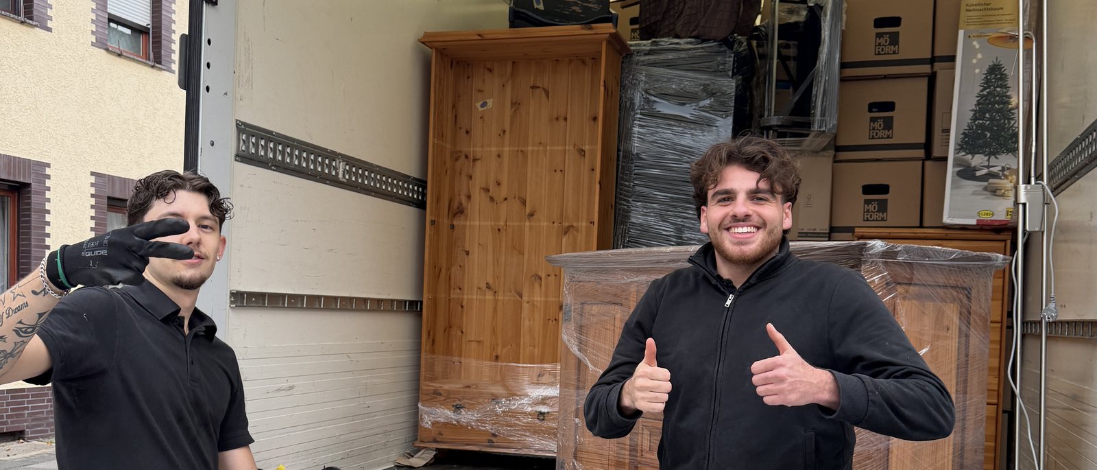 Umzugsteam für Bornheim: Zwei Helfer in blauer Arbeitshose vor einem gepackten Umzugswagen, einer mit Daumen hoch