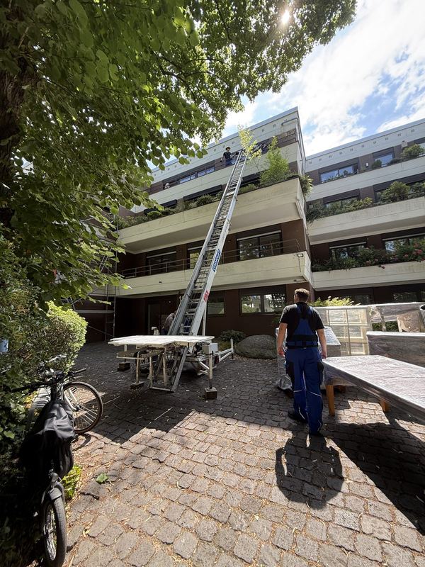 An einem mehrstöckigen Wohnhaus steht ein ausgefahrener Möbellift, unten wartet ein Kollege