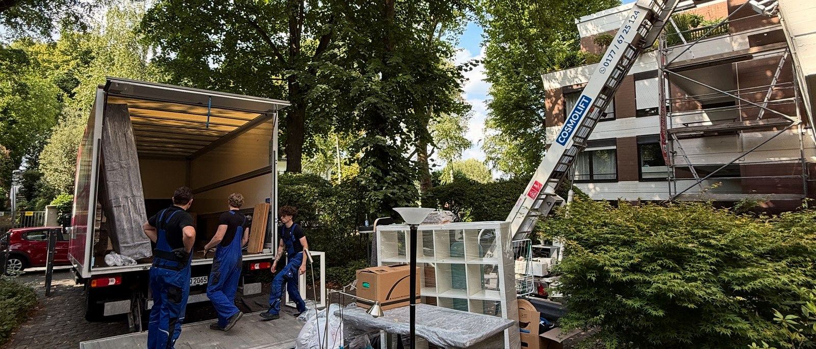 Möbellift für Umzüge in Bornheim: Ein aufgestellter Möbellift führt bis in das obere Stockwerk eines Wohnhauses mit Gerüst, davor stehen drei Umzugshelfer an einem geöffneten Lkw-Laderaum neben einem Regal und Kartons