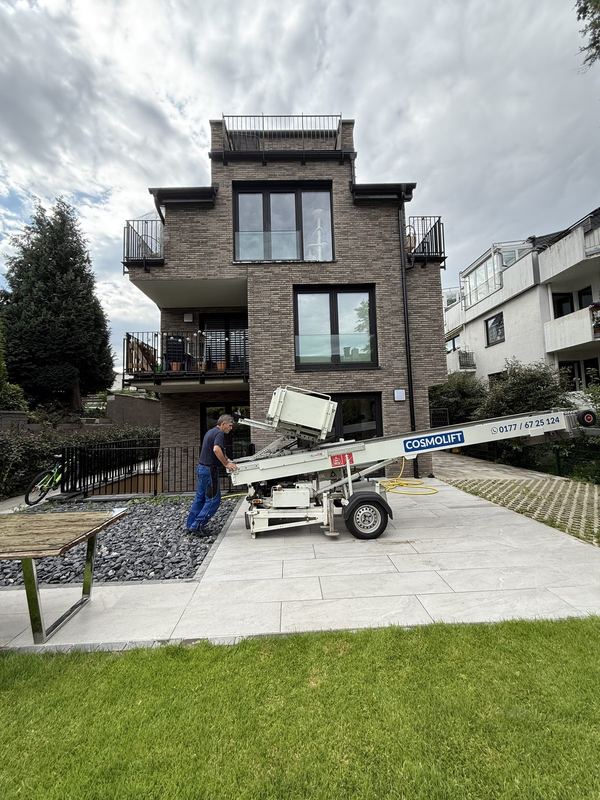 Vor einem modernen Klinkerbau mit Balkonen steht ein Mitarbeiter neben einem Möbellift, der noch eingeklappt auf einem Anhänger liegt