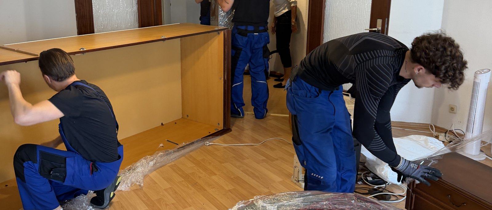 Möbelmontage in Bornheim: Drei Mitarbeiter zerlegen in einem Wohnzimmer mit Parkett eine große Schrankwand aus Holz, die Seitenteile liegen bereits am Boden, daneben Folie und Werkzeug.