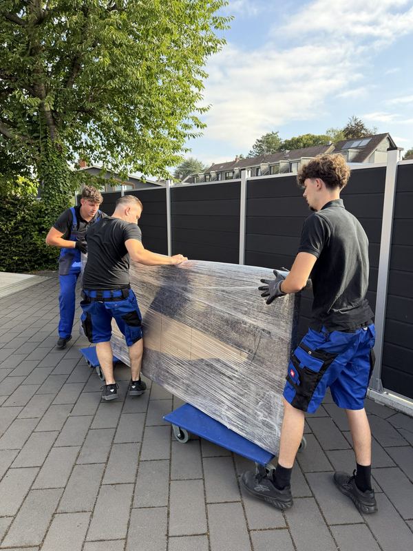 Über eine Einfahrt schieben drei Kollegen ein in Folie gepacktes Möbelstück auf Rollbrettern