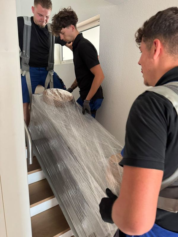 Zu dritt tragen Kollegen ein in Folie gewickeltes Möbelstück die Treppe hinab