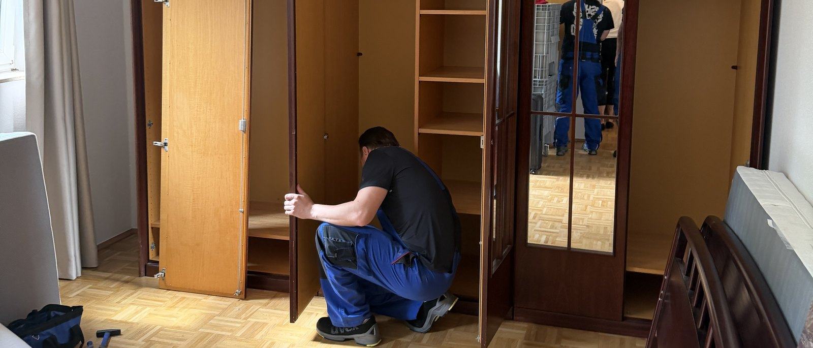 Seniorenumzug in Bornheim: Ein Umzugshelfer kniet in einem Schlafzimmer mit Parkett vor einem ausgeräumten Schrank aus dunklem Holz