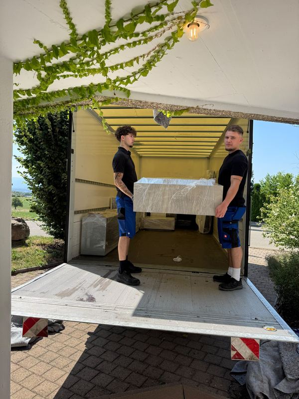 Zwei Umzugshelfer hieven ein folienverpacktes Möbelstück über die Ladebordwand hinauf in den Wagen.