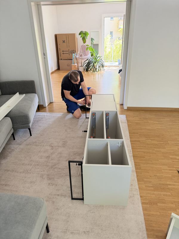 In einem hellen Wohnzimmer kniet ein Umzugshelfer vor einem weißen Sideboard und baut es zusammen, hinter ihm stehen Umzugskartons.