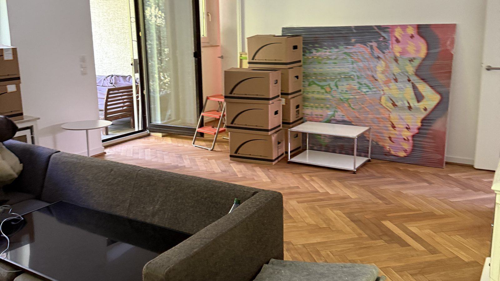 Umzugscheckliste für Bornheim: In klaren Schritten ins neue Zuhause: In einem Wohnzimmer mit Fischgrätparkett stehen gestapelte Umzugskartons neben einem Sofa und einem Beistelltisch, an der Wand lehnt ein großes buntes Bild, dahinter eine Terrassentür.