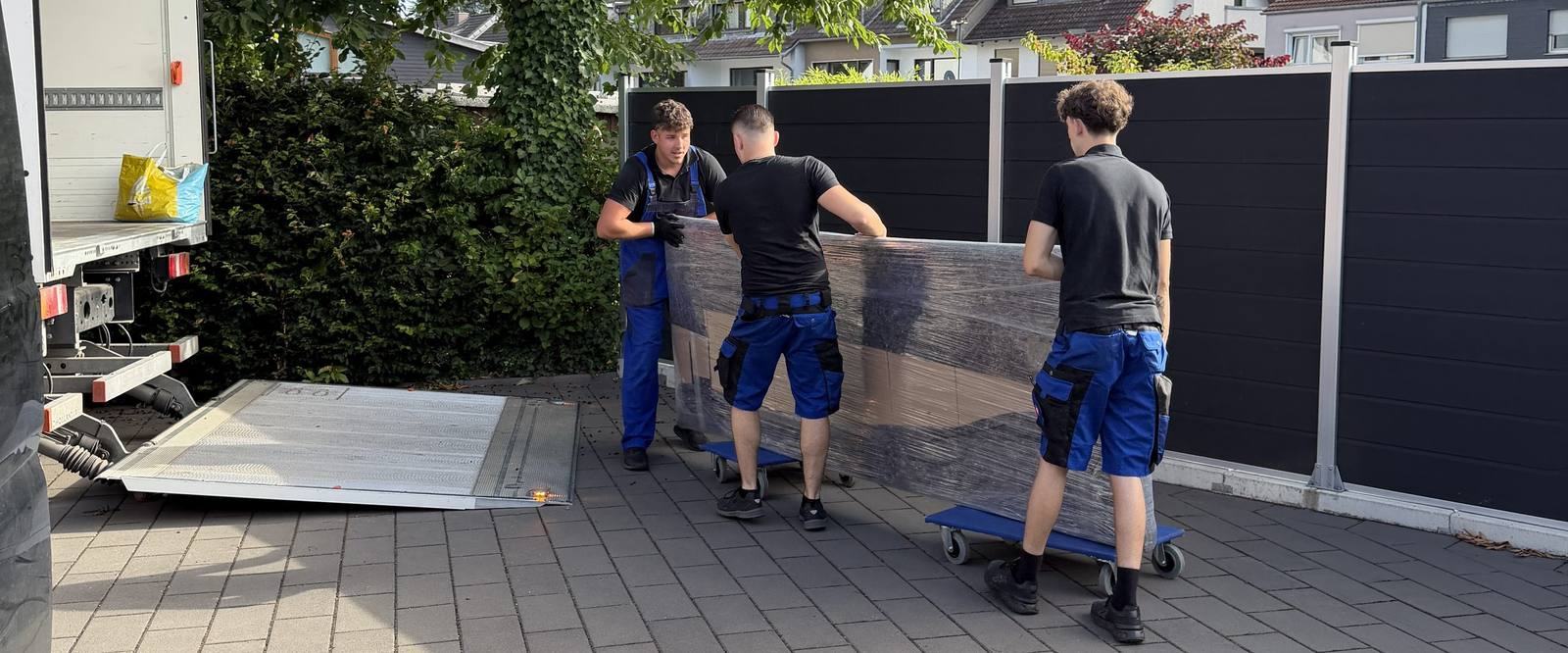 Umzugsunternehmen Bornheim: Drei Umzugshelfer schieben ein folienverpacktes Möbelstück auf Rollbrettern zu einem Umzugswagen, dessen Ladebordwand abgesenkt ist.