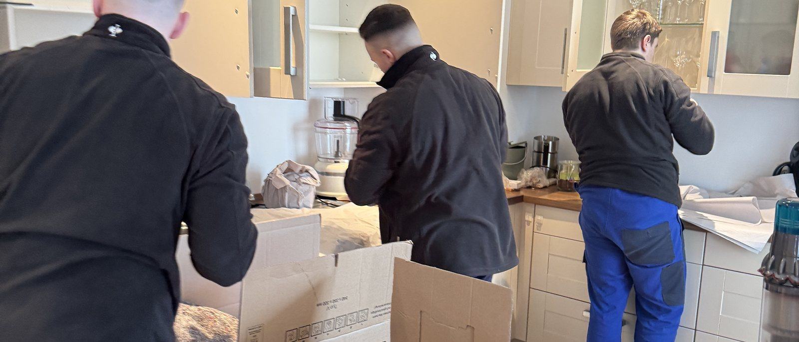 Packen für den Umzug in Bornheim: Kartons und mehr: Drei Mitarbeiter packen in einer Küche mit hellen Fronten Geschirr und Kleingeräte in Umzugskartons auf der Arbeitsplatte, in der Mitte ein offener Karton mit Packpapier.