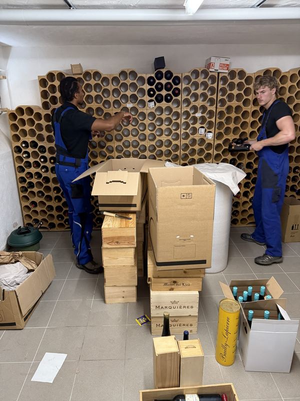 Zwei Umzugshelfer holen Weinflaschen aus einem Kellerregal aus Tonröhren und setzen sie in Holzkisten und Kartons.
