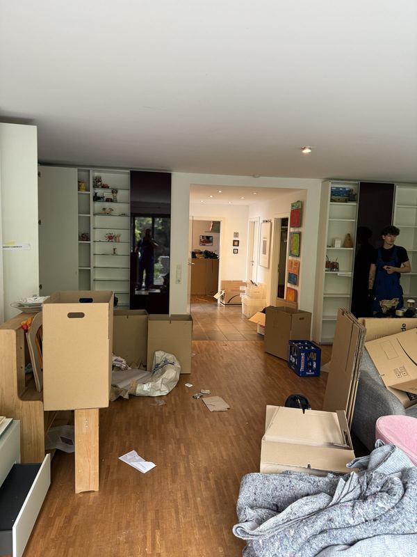 Ein Wohnzimmer mit schon leeren Einbauregalen und bereitgestellten Kartons, hinten zwei Kollegen