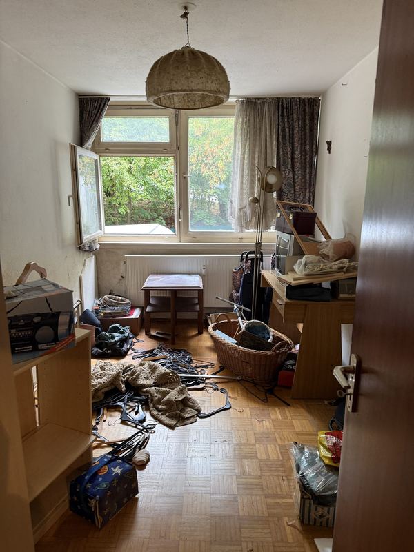 Vollgestelltes Zimmer vor dem Ausräumen Ein dicht vollgestelltes Zimmer mit Parkett, auf dem Boden liegen Körbe, Taschen, Kabel und Hausrat, hinten ein Fenster, durch das man ins Grüne sieht
