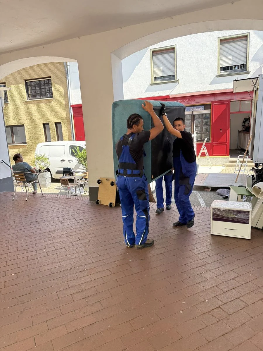 Zwei Umzugshelfer tragen eine Tafel unter einer historischen Arkade hindurch